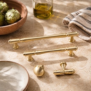 May include: A collection of brushed gold hardware including two long cabinet pulls with ribbed detailing, one round knob, and one T-shaped handle. The metal pieces feature a smooth, matte finish and cylindrical shapes with decorative grooved accents.