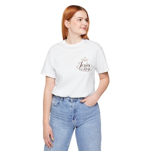 May include: A white short-sleeved t-shirt featuring a small graphic on the upper left chest. The graphic consists of a gold-colored crown icon positioned above the brown cursive text that reads Jesús es Rey.