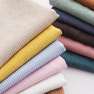 Può includere: Una pila di tessuti in velluto a coste piegati in vari colori, tra cui beige, giallo, azzurro, rosa, bianco, marrone, blu scuro, verde salvia e arancione ruggine. Ogni pezzo di tessuto presenta una distinta trama a coste verticali sulla superficie.