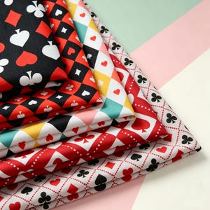 Peut inclure: Une pile de tissus pliés présentant divers motifs de cartes à jouer. Les designs incluent des carreaux, cœurs, trèfles et piques noirs et rouges disposés en grilles géométriques, avec des sections affichant des losanges multicolores sur fond blanc.