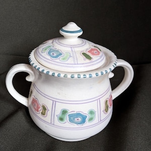May include: A white ceramic sugar bowl with two handles and a matching lid. The bowl features a hand-painted pattern of light purple rectangular borders containing stylized blue, pink, and green floral motifs, with a dotted blue rim.