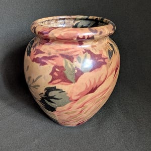 May include: A rounded ceramic vase featuring a smooth, glossy finish with a multi-coloured floral pattern in shades of peach, muted red, dark green, and black. The vase has a wide, circular opening with a thick, rolled rim and a tapered base.