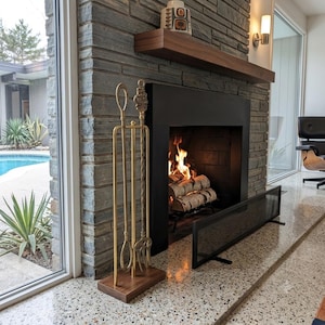 May include: A brass fireplace tool set featuring a poker, shovel, and tongs with decorative handles, mounted on a rectangular walnut wood base. The set stands on a speckled stone hearth next to a black metal fireplace screen and a burning fire.