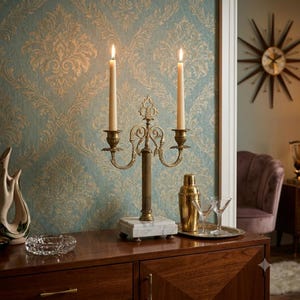 May include: A brass candelabra with two lit taper candles, featuring an ornate central stem and curved arms, mounted on a square white marble base. The metal fixture has a metallic gold finish and intricate scrollwork details on the arms.