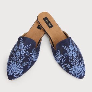 Könnte beinhalten: Ein Paar dunkelblaue bestickte Hausschuhe mit spitzer Zehenpartie. Die Hausschuhe haben ein florales Design in einem helleren Blauton. Der Markenname "SAACHI" ist auf der Innensohle aufgedruckt.