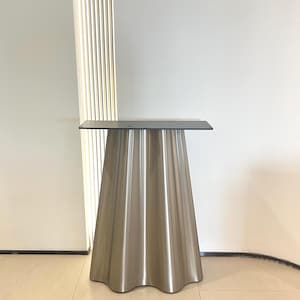 May include: A metallic table base with a conical, fluted design featuring vertical ridges. The base is topped with a flat, rectangular black mounting plate that has pre-drilled holes for attachment, providing a stable support structure.