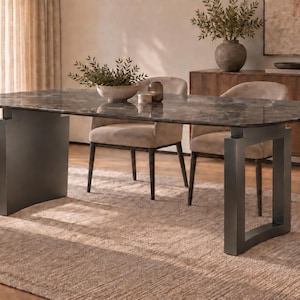 Puede incluir: Una mesa de comedor rectangular con una superficie de mármol gris oscuro pulido y una base de metal oscuro resistente con dos patas anchas en forma de U. La mesa está rodeada por cuatro sillas tapizadas en tela de color beige claro.