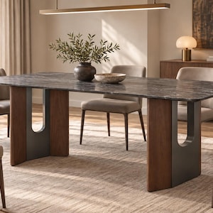 Puede incluir: Una mesa de comedor rectangular con una superficie de mármol gris oscuro y una base con dos soportes en forma de U hechos de madera oscura y metal gris mate. La mesa está rodeada por cuatro sillas tapizadas en tela beige claro con patas de madera.