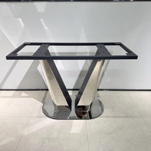 Puede incluir: Una base de mesa con un marco rectangular de metal negro sostenido por dos pilares en forma de V con acabado plateado pulido. La estructura descansa sobre una base ovalada plana y reflectante, proporcionando un soporte estable.