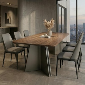 Puede incluir: Una mesa de comedor rectangular con una superficie de madera oscura y una base gris sólida y angular, rodeada de seis sillas tapizadas en gris. Un jarrón de cerámica claro con hierba de la pampa seca se encuentra sobre la mesa con libros.