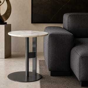 Può includere: Un tavolino rotondo con piano in marmo bianco caratterizzato da un sottile motivo a venature grigie. Il tavolo è sostenuto da una colonna centrale in metallo grigio scuro collegata a una base circolare piatta con la stessa finitura.