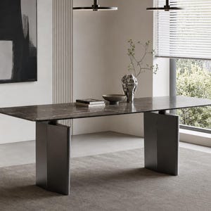 Può includere: Un tavolo da pranzo rettangolare con piano in marmo marrone scuro venato di bianco, sostenuto da due spesse gambe verticali in metallo grigio antracite. Il piano del tavolo ospita una pila di libri, una ciotola bassa e un vaso metallico.
