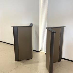 Puede incluir: Dos patas de mesa en forma de L con un marco metálico oscuro y un panel central liso de color marrón claro. Cada pata tiene una placa de montaje negra plana en la parte superior con orificios pretaladrados, sobre un suelo claro.