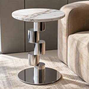 Pode incluir: Uma mesa lateral redonda com tampo de mármore branco com veios cinzentos. A base consiste numa coluna vertical de cinco segmentos cilíndricos de cromo polido, apoiada numa placa de base circular e refletora de cromo no chão.