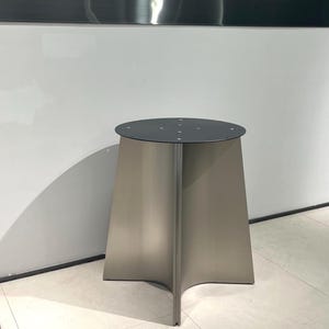 May include: A tall, freestanding pedestal table featuring a circular, matte black top with visible screw points. The base is constructed from three curved, metallic silver panels that meet in the centre, creating a flared, tripod-like structure.