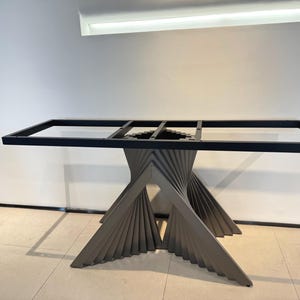 Puede incluir: Una base de mesa con un marco rectangular de metal negro sostenido por un pedestal central. El pedestal está construido con múltiples listones grises inclinados dispuestos en un patrón de abanico en espiral que converge al centro.