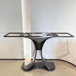 Puede incluir: Una mesa consola de metal oscuro con una parte superior de marco abierto rectangular sostenida por una base de pedestal central. La base consta de dos soportes curvos y simétricos que se ensanchan en la parte inferior.