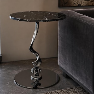 May include: A round side table featuring a black marble top with white veining. The table is supported by a single, vertically twisted, polished silver-toned metal pedestal base that connects to a flat, circular, reflective black metal floor plate.