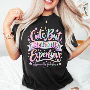 Może przedstawiać: Czarny t-shirt z kolorowym nadrukiem graficznym w pastelowych odcieniach, zawierający napis "Cute But Medically Expensive" oraz "chronically fabulous" stylizowaną czcionką, ozdobiony małymi gwiazdkami, sercami i ikonami kwiatów.