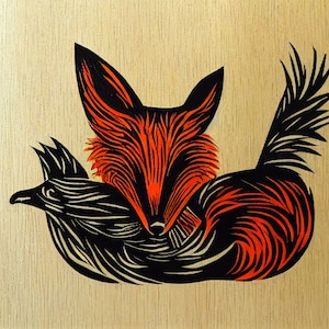 May include: A black and orange woodblock print of a fox with a bird in its mouth. The fox is facing forward and the bird is facing to the left. The print is on a light wood background.