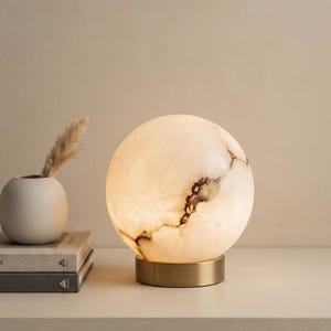 May include: A spherical table lamp featuring a translucent, cream-colored stone shade with natural brown veining, illuminated from within. The lamp rests on a circular, brushed brass-toned metal base, providing a stable foundation for the globe.