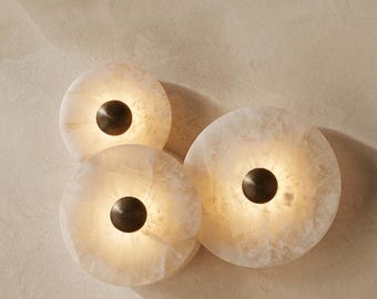 May include: Three circular wall lights featuring translucent, off-white stone discs with natural veining. Each light fitting has a central, dark bronze-toned metal cap with a concentric, ribbed, circular pattern that emits a warm, soft glow.