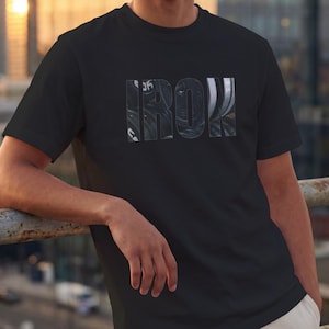 Könnte beinhalten: Ein schwarzes T-Shirt mit kurzen Ärmeln und dem Wort IRON in fetter Blockschrift auf der Brust. Die Buchstaben sind mit einer fotografischen Grafik von gestapelten Metall-Hantelscheiben gefüllt, die ein dunkles, strukturiertes Finish zeigen.