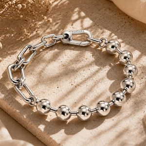 Puede incluir: Una pulsera de tono plateado que combina cuentas metálicas pulidas y eslabones rectangulares. La pieza incluye un cierre de mosquetón funcional grabado con la palabra PANDORA, que conecta las dos secciones distintas de la pulsera.