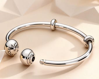 May include: A polished silver-tone open bangle bracelet featuring two spherical end caps engraved with the word PANDORA. The bracelet includes two additional sliding spherical beads along the smooth, curved metal band, which has a small notched clasp.