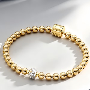 May include: A gold-toned beaded bracelet featuring a series of polished spherical beads. The design includes a single textured, silver-toned bead encrusted with small clear stones and a rectangular gold-toned clasp with engraved markings.