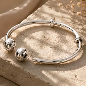 Può includere: Un bracciale rigido aperto in argento lucido con una fascia circolare liscia. Il bracciale termina con due ciondoli sferici, uno dei quali è rimovibile, e include una perlina scorrevole più piccola. La parola PANDORA è incisa sui ciondoli.