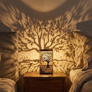 Può includere: Una lampada cilindrica in metallo con un esterno scuro caratterizzato da un design ad albero della vita tagliato al laser. Quando è accesa, la lampada proietta sulla parete retrostante una grande e dettagliata ombra di un albero.