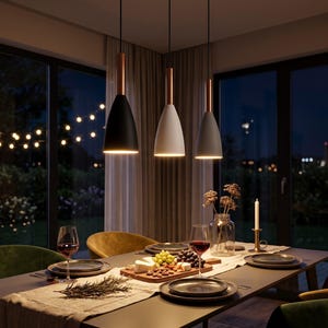 Puede incluir: Tres lámparas colgantes sobre una mesa de comedor. Cada lámpara tiene una pantalla en forma de campana de color negro, blanco o gris, rematada con una pieza cilíndrica de color cobre. Se suspenden mediante cables negros delgados.