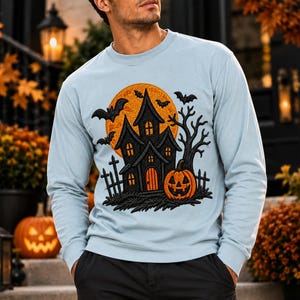 May include: A light blue long-sleeve sweatshirt featuring a graphic print of a dark haunted house, a large orange full moon, flying bats, a bare tree, and a carved jack-o'-lantern pumpkin with a fence in the foreground.