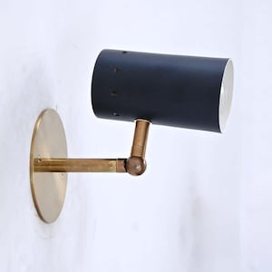 Op de afbeelding: Een wandlamp met een cilindervormige, matzwarte kap voorzien van kleine ventilatiegaatjes en een witte binnenkant. Het armatuur heeft een messingkleurige ronde muurplaat en een verstelbare horizontale arm met een draaipunt.