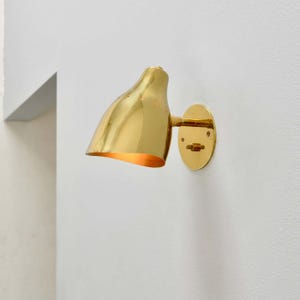 May include: A polished brass wall sconce featuring a curved, bell-shaped shade with a series of small perforations along the top edge. The fixture is mounted to a circular brass backplate and extends outward with a short, adjustable arm.