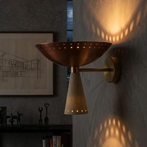 Op de afbeelding: Een wandlamp met een brede, koperkleurige komvormige kap met een geperforeerde rand en een naar beneden gerichte, getextureerde, gebroken witte conische voet. Het armatuur verspreidt licht door de perforaties en werpt patronen op de muur.