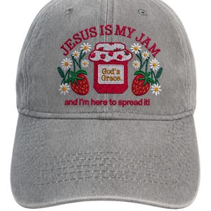Könnte beinhalten: Eine graue Baseballkappe mit gesticktem Text, der JESUS IS MY JAM und and I'm here to spread it! lautet. Das Design zeigt ein rotes Glas mit der Aufschrift God's Grace, umgeben von roten Erdbeeren und weißen Gänseblümchen mit grünen Stielen.