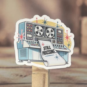 May include: A die-cut sticker featuring a light blue vintage mainframe computer with two reel-to-reel tapes. A paper strip feeds out from the front with the text STILL PROCESSING... printed in black. The design includes red and blue starburst accents.