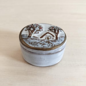 Peut inclure: Une petite boîte à bijoux ronde en céramique avec un vernis blanc et un bord métallique brun. Le couvercle présente une scène peinte à la main représentant une maison nichée entre deux arbres avec de petites touches rouges, en tons brun et blanc.