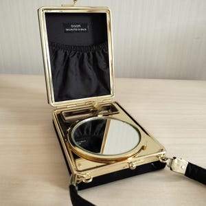 May include: A black velvet evening bag with a gold-tone metal frame and clasp. The interior features a black fabric lining with a gathered pocket and a label reading SILVER ORIGINATED IN SPAIN. A round mirror is mounted on the lower interior section.
