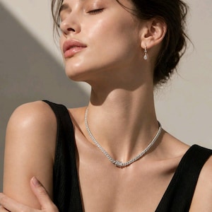 May include: A silver-toned curb chain necklace featuring a rectangular clasp with embedded clear stones, paired with a matching silver-toned earring that has a small, dangling, pear-shaped clear stone drop. The jewelry is metallic and reflective.