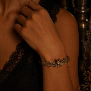 Può includere: Un bracciale in metallo color oro con un design dettagliato di un pipistrello con le ali spiegate. Il pipistrello è posizionato al centro del cinturino, che presenta una superficie testurizzata e una finitura scura in stile antico sulle ali.