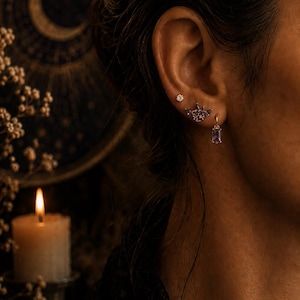 Peut inclure: Un ensemble de trois boucles d'oreilles dorées portées à une oreille. La boucle supérieure est un petit clou rond, celle du milieu est en forme d'ailes avec des pierres violettes, et celle du bas est un anneau doré avec une pierre violette rectangulaire.
