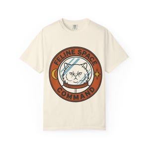 Könnte beinhalten: Ein cremefarbenes Kurzarm-T-Shirt mit einer kreisförmigen Grafik auf der Brust. Das Design zeigt eine Katze in einem Weltraumhelm mit dem Text FELINE SPACE COMMAND in einem dunkelorangen Rand, ergänzt durch einen kleinen gelben Halbmond und einen Stern.
