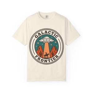 May include: An off-white short-sleeve t-shirt featuring a circular graphic with a rope border. The design shows a desert landscape with cacti, a cowboy on horseback, and a UFO beaming light down, with the text GALACTIC FRONTIER in dark lettering.
