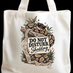 Puede incluir: Una bolsa de tela blanca con el gráfico de una serpiente con gafas de sol enroscada en un letrero de madera que dice DO NOT DISTURB. Shedding. El diseño incluye follaje verde, una piel de serpiente mudada y pequeños detalles de estrellas.
