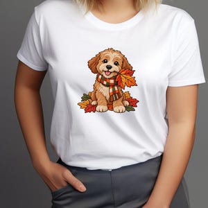 Puede incluir: Una camiseta blanca de manga corta con un estampado gráfico de un cachorro de color marrón claro que lleva una bufanda de cuadros. El cachorro está sentado entre hojas de otoño de colores y sostiene una hoja de arce naranja en la boca.
