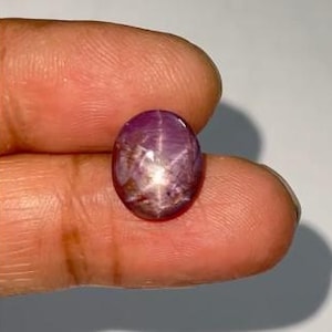 Könnte beinhalten: Ein polierter, ovaler violetter Sternsaphir-Edelstein mit einem ausgeprägten sechsstrahligen Sterneffekt, bekannt als Asterismus. Der im Cabochon-Schliff gearbeitete Stein zeigt einen tiefvioletten Farbton mit einer hellen, zentrierten Lichtreflexion.