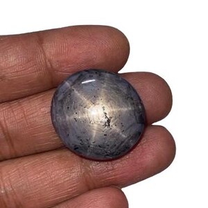 May include: A round, cabochon-cut star sapphire gemstone with a greyish-blue hue. The stone features a distinct six-rayed asterism effect, where a bright, centred star pattern reflects light across the polished, smooth, and slightly translucent surface.